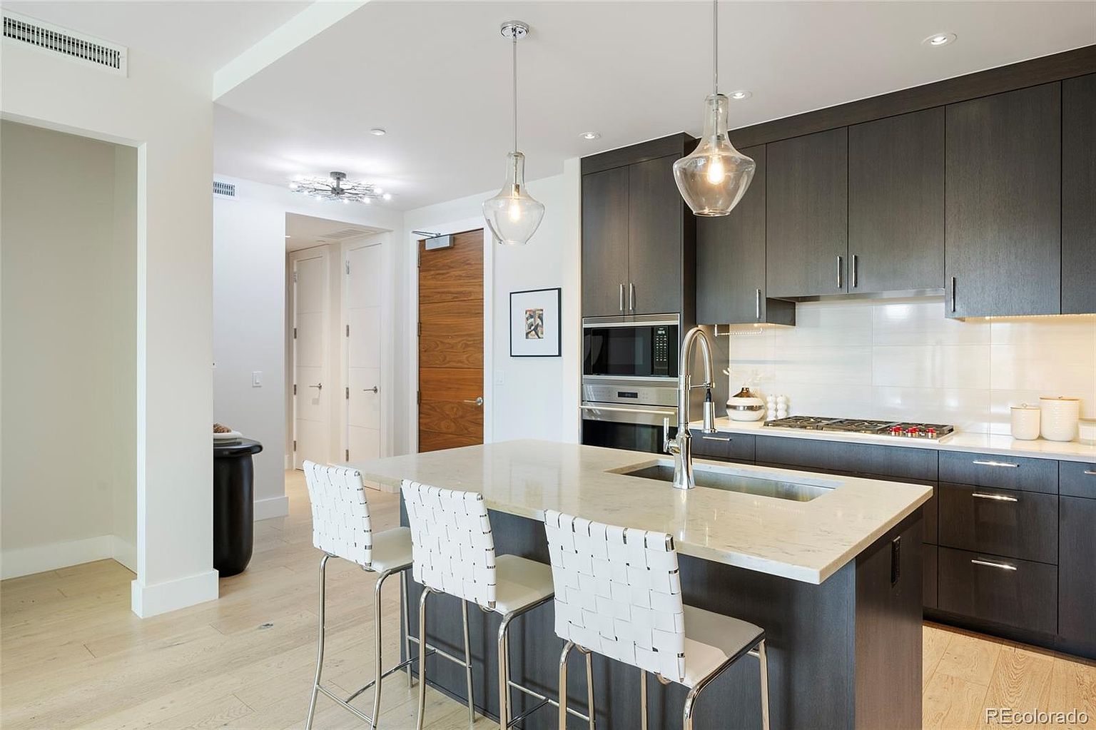 This is a modern kitchen featuring dark wood cabinetry, stainless steel appliances, and a light-colored countertop island with three bar stools. Pendant lights hang above the island, and the backsplash is a light tile. The perspective is from the front, showcasing the island and the kitchen's layout.