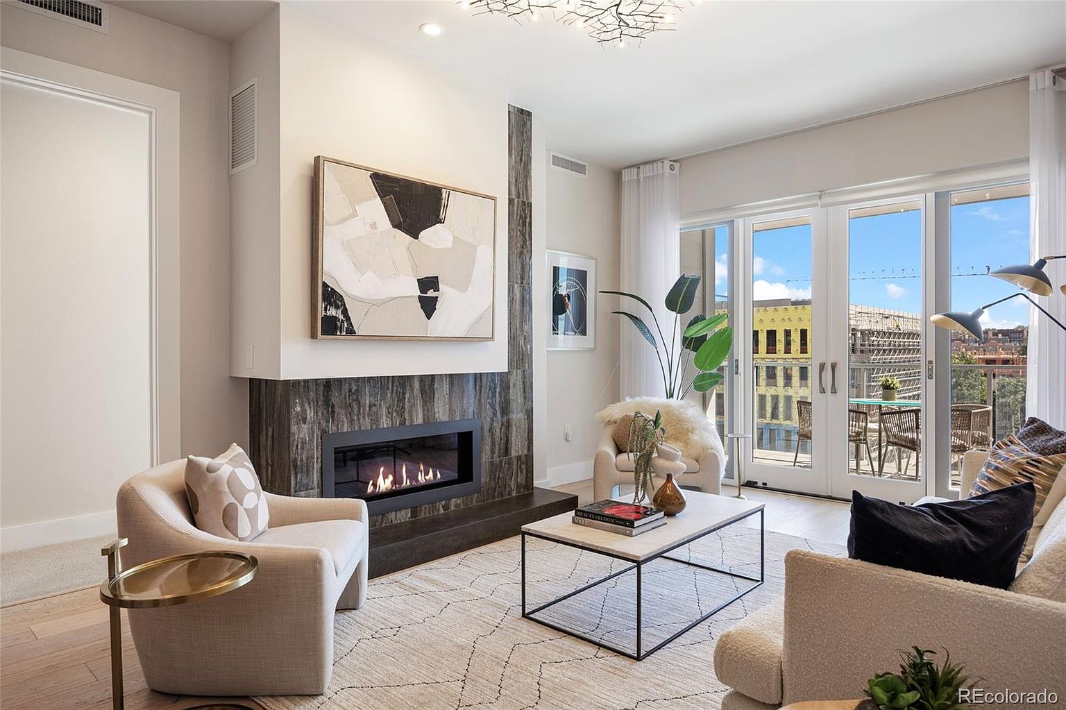 This is a bright and stylish living room featuring a modern fireplace with a large abstract art piece above it. The room is furnished with comfortable seating, including armchairs and a sofa, arranged around a marble-topped coffee table. Large glass doors lead to a balcony, offering a view of the cityscape and filling the room with natural light.