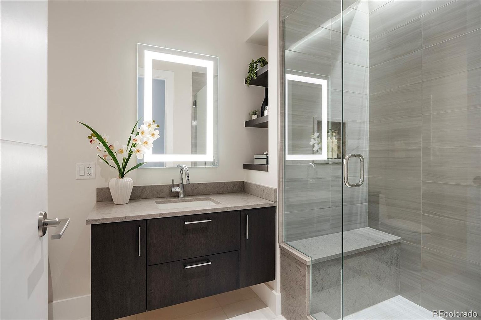 This is a modern primary bathroom featuring a dark wood vanity with a light countertop and a square, illuminated mirror. A glass-enclosed shower with gray tiling and a built-in bench is visible to the right. The overall impression is clean, contemporary, and luxurious.