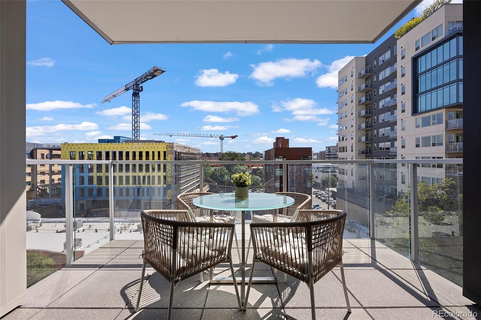 This image showcases a stylish balcony with a modern aesthetic. The balcony features a glass-topped round table surrounded by four woven chairs with cushions, creating an inviting outdoor seating area. The view from the balcony includes city buildings and construction cranes under a partly cloudy sky, offering an urban backdrop.