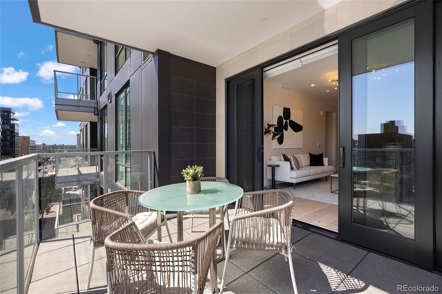 This image showcases a stylish balcony featuring a round glass-top table surrounded by four woven chairs with cushions, creating an inviting outdoor dining or relaxation space. The balcony offers views of the city skyline, and the sliding glass doors seamlessly connect the indoor living area to the outdoor space. The modern design and comfortable seating make this balcony an appealing extension of the home.