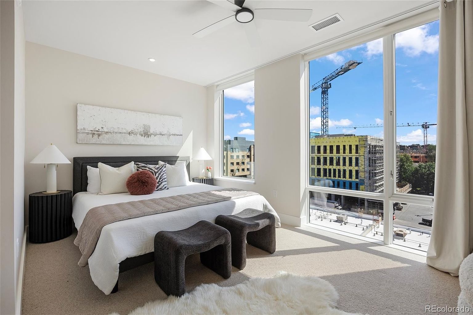 This is a bright and airy primary bedroom featuring a large window with city views, a comfortable bed with stylish pillows and a throw, and modern bedside tables with lamps. The room is decorated in neutral tones, creating a serene and inviting atmosphere. Two small benches are placed at the foot of the bed, and a fluffy rug adds texture to the space.
