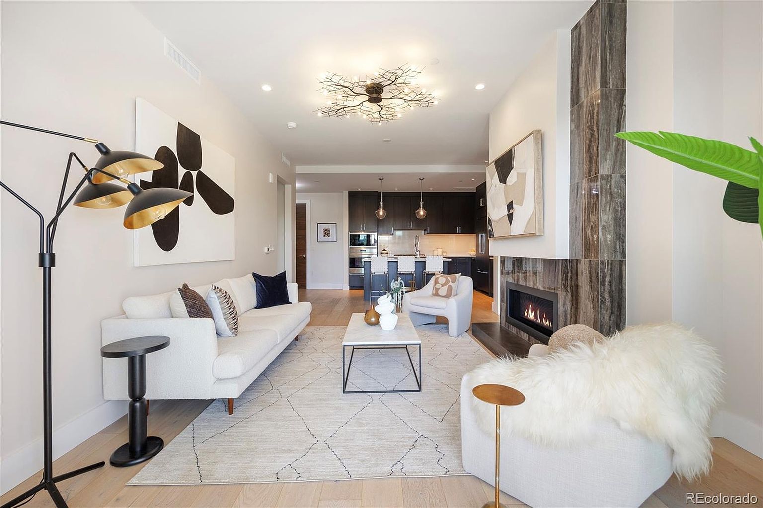 This is an interior shot of a modern living room, showcasing a bright and airy space with a white sofa, a stylish black floor lamp, and a unique ceiling light fixture. The room features a fireplace with a striking stone surround, complemented by abstract artwork and a neutral-toned rug, creating a sophisticated and inviting atmosphere.