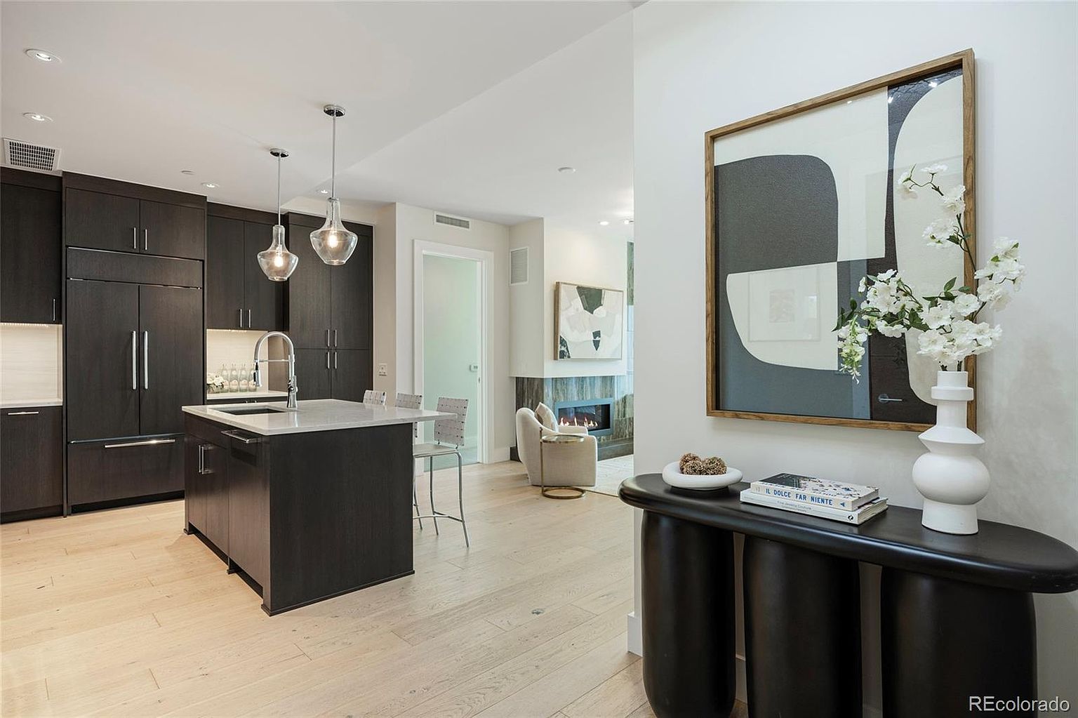 This interior shot showcases a modern kitchen with dark wood cabinetry and a light countertop island. Pendant lights hang above the island, and stainless steel appliances are integrated into the cabinetry. The space flows into a living area with a fireplace, creating an open and inviting atmosphere.