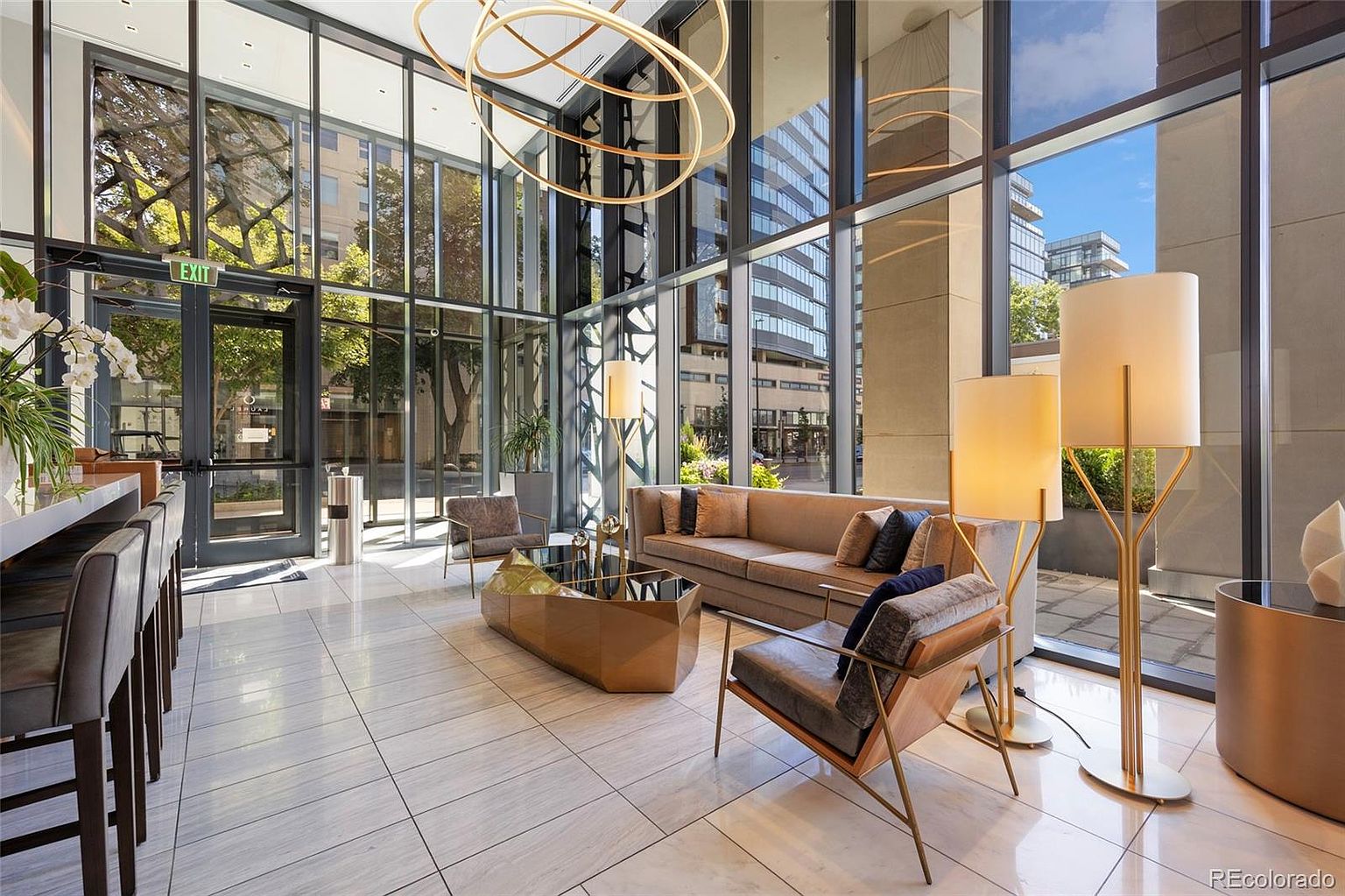 This is a bright and modern lobby area featuring floor-to-ceiling windows that offer ample natural light and views of the surrounding cityscape. The space is furnished with contemporary seating, including a sofa and armchairs, arranged around a geometric coffee table. A striking circular chandelier hangs from the high ceiling, adding a touch of elegance.