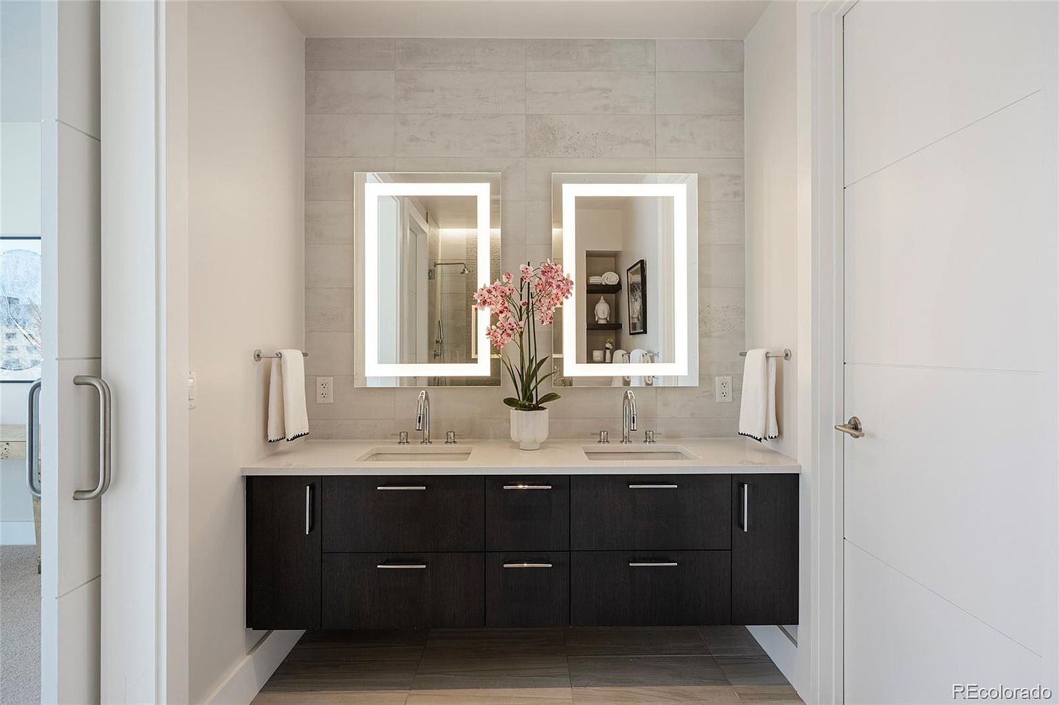 This is a well-lit primary bathroom featuring a double vanity with a dark wood cabinet and a white countertop. Two illuminated rectangular mirrors hang above the sinks, complemented by a light gray tiled backsplash. A decorative floral arrangement sits between the sinks, adding a touch of elegance to the modern design.