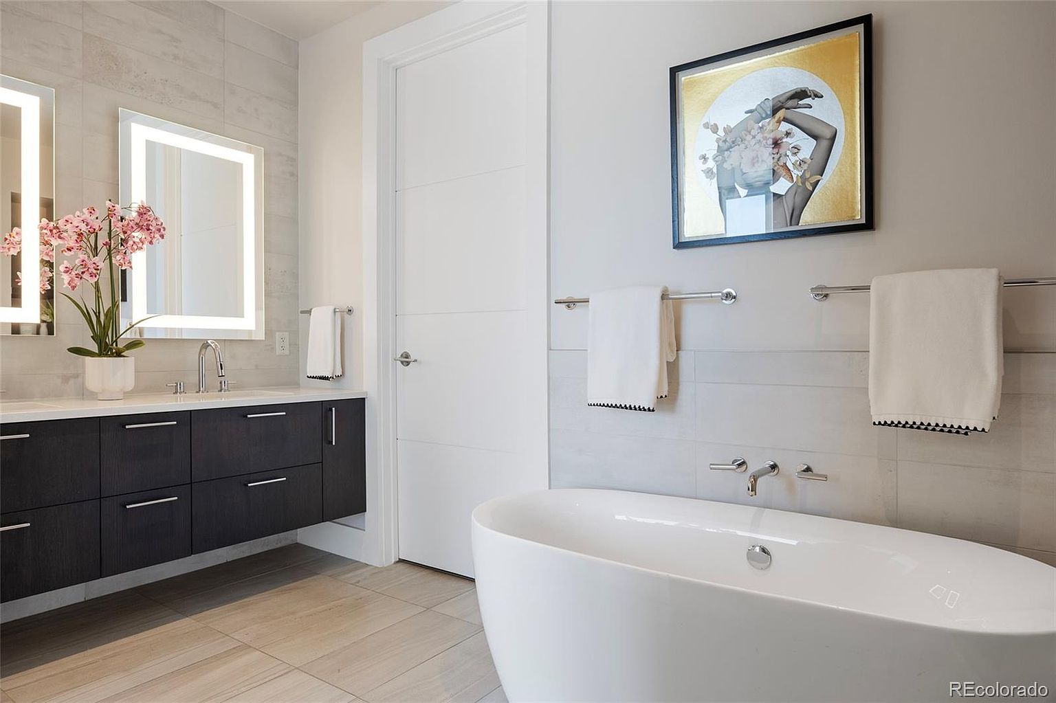 This is a modern primary bathroom featuring a freestanding white bathtub, a floating dark wood vanity with dual sinks, and illuminated mirrors. The walls are tiled in a light gray, and a piece of art hangs above the tub, adding a touch of elegance. The overall impression is clean, luxurious, and well-designed.