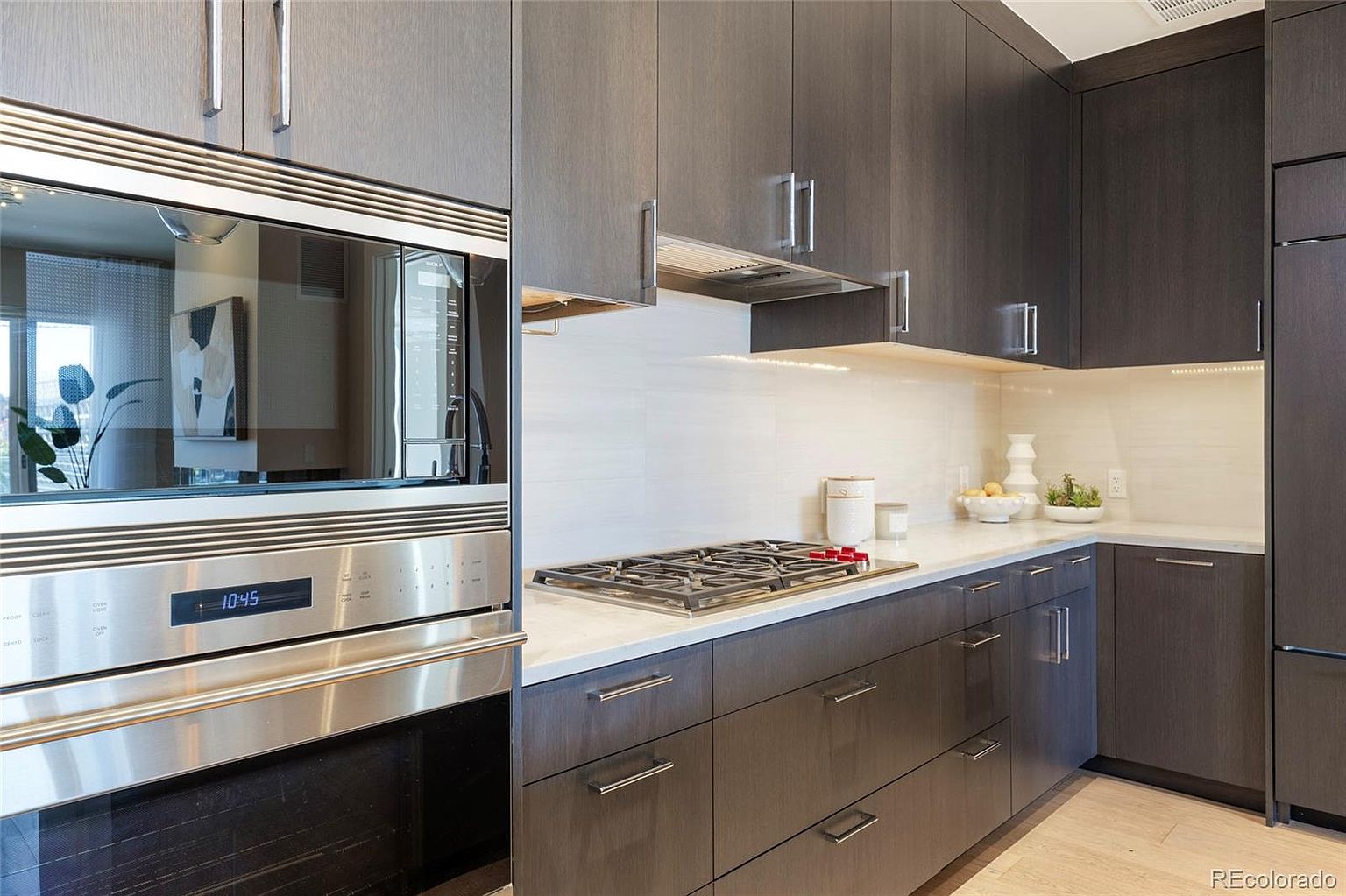 This is a modern kitchen featuring dark wood cabinetry with sleek, silver hardware. Stainless steel appliances, including a built-in microwave and oven, add a contemporary touch. The countertops are light-colored, contrasting with the dark cabinets, and the backsplash is a clean, white tile.