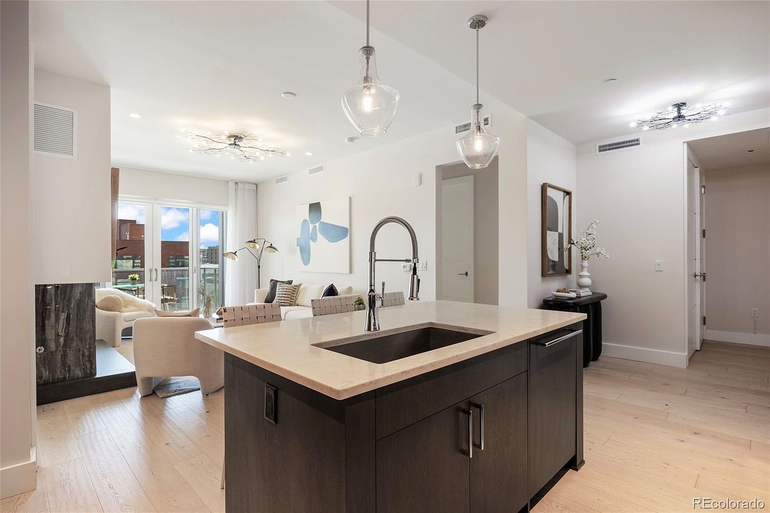 This is an interior shot of a modern kitchen that flows into a living area. The kitchen features a dark wood island with a light countertop and a stainless steel sink and faucet. The living area has a fireplace, a sofa, and access to a balcony, creating a bright and airy living space.