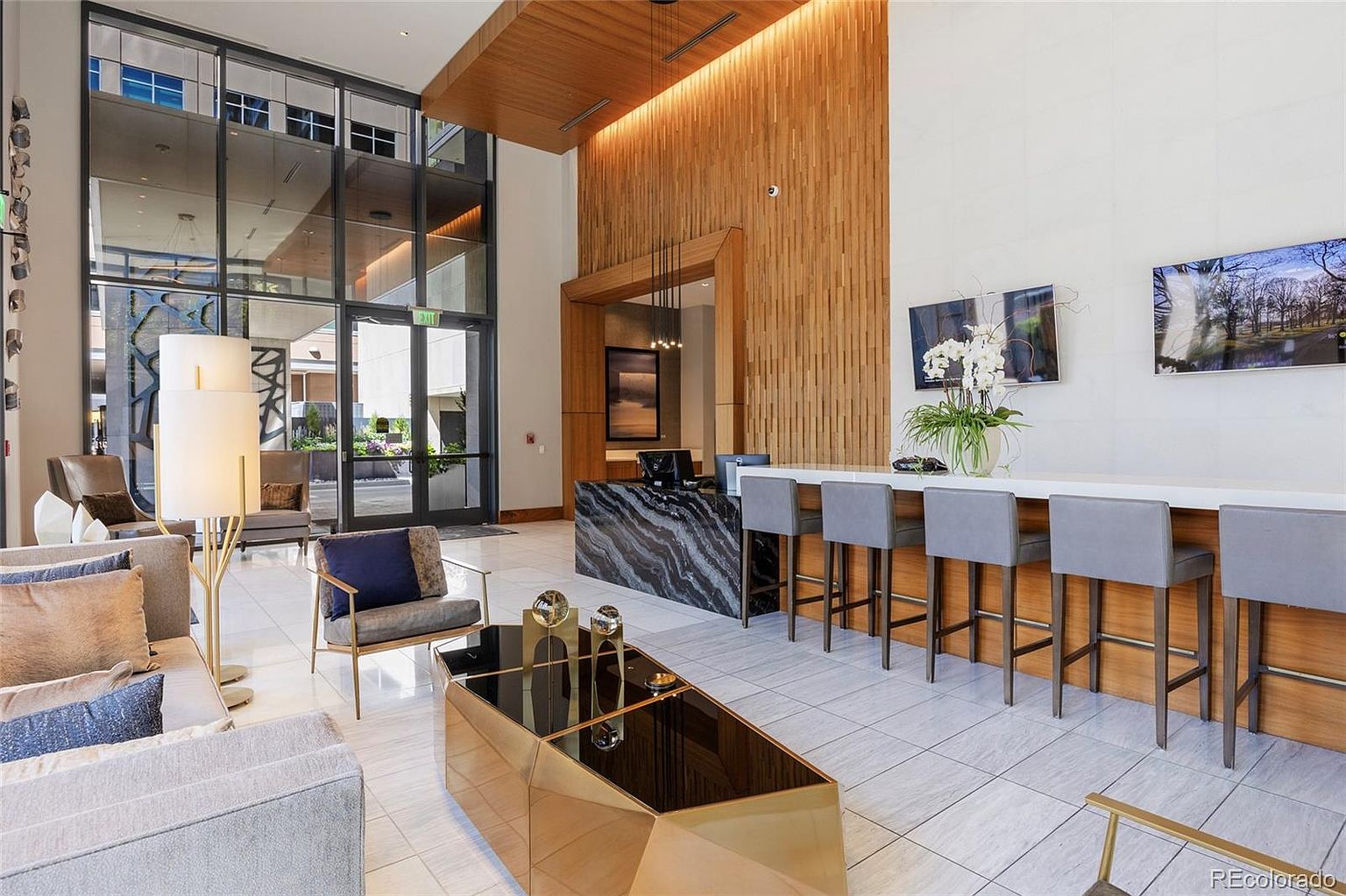 This is an interior shot of a modern lobby or hallway area. The space features a reception desk with a marble countertop, a seating area with contemporary furniture, and a high ceiling with wood paneling. The overall impression is luxurious and inviting, suggesting a high-end residential building or hotel.