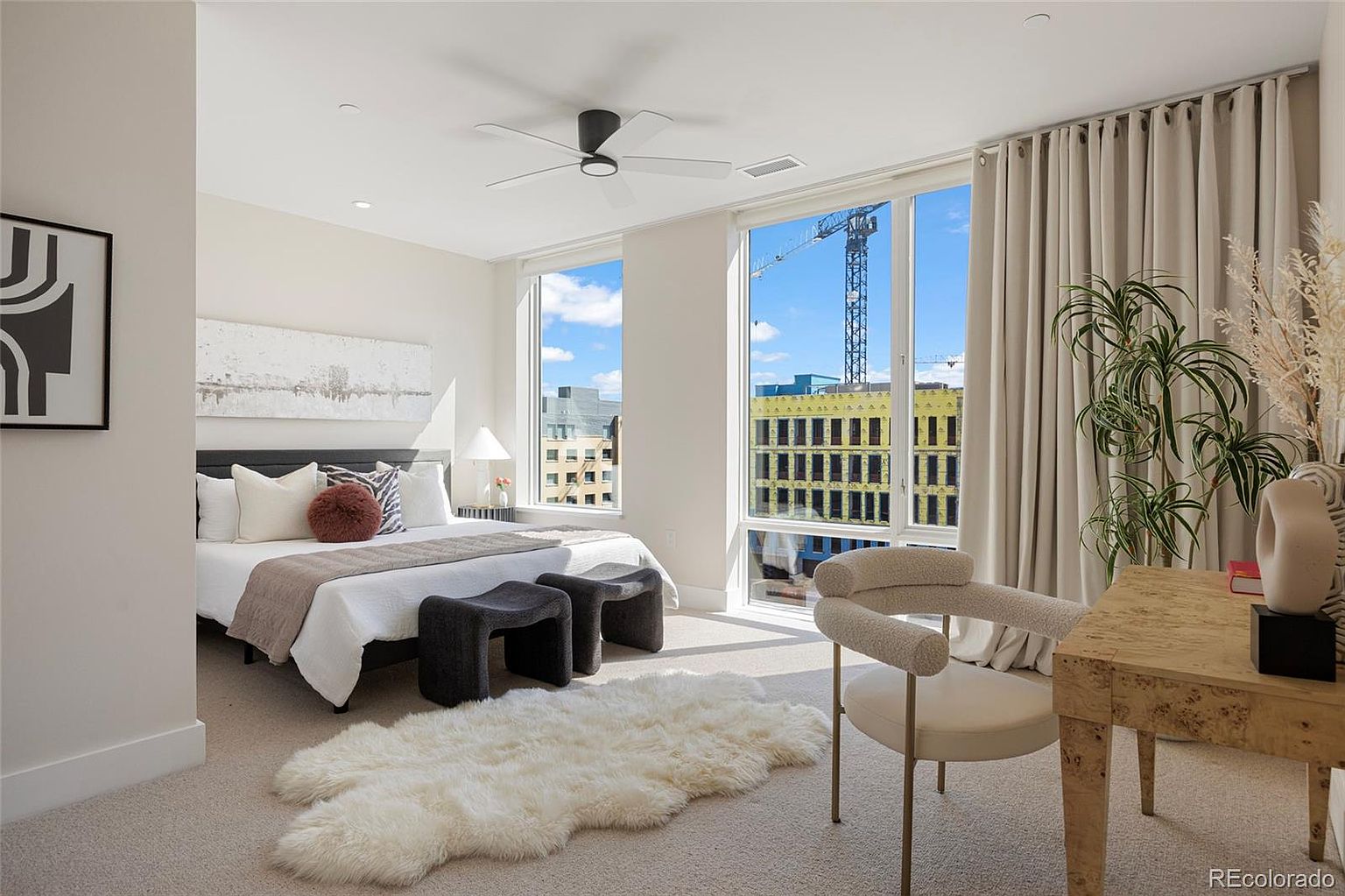 This is a bright and airy primary bedroom featuring a large bed with neutral bedding, complemented by modern furniture and decor. Large windows offer city views, enhanced by light-filtering curtains. The room is well-lit and styled with a contemporary aesthetic, creating a serene and inviting atmosphere.