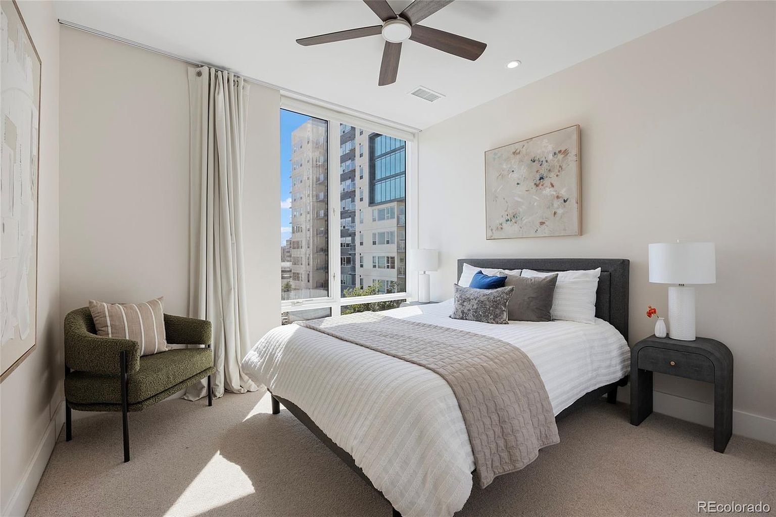 This is a well-lit primary bedroom featuring a neutral color palette. The room includes a bed with a gray headboard, white bedding, and decorative pillows. A large window provides natural light and a view of the city, complemented by light-colored curtains and a comfortable armchair. A ceiling fan and a modern nightstand with a lamp add to the room's appeal.