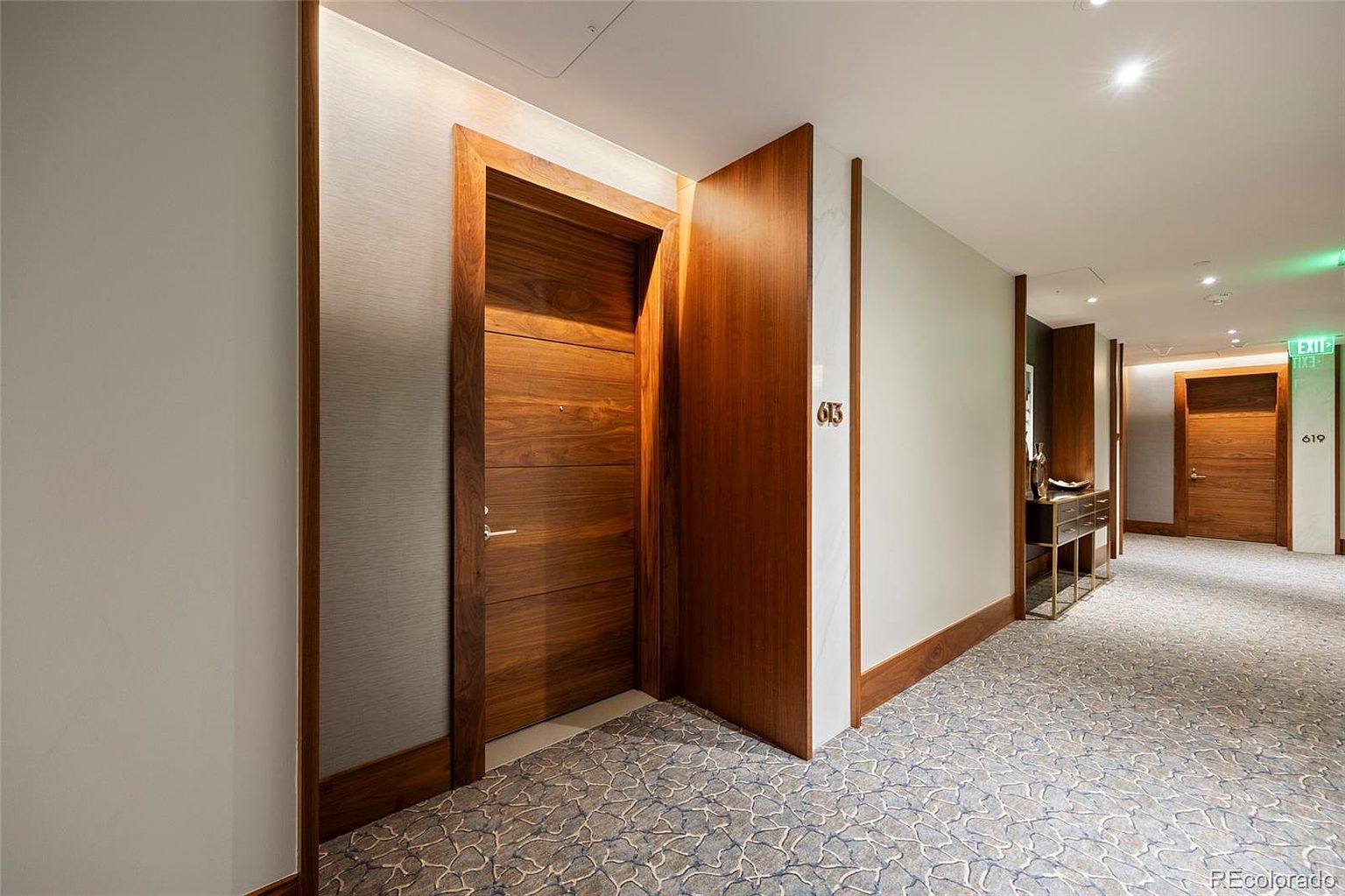This interior shot showcases a well-lit hallway with a patterned carpet and wooden accents. The hallway features doors with room numbers, suggesting an apartment or hotel setting. A console table with decorative items adds a touch of elegance to the space, creating a welcoming and sophisticated atmosphere.
