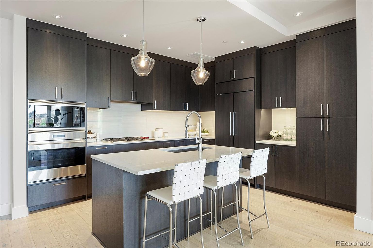 This is a modern kitchen featuring dark wood cabinetry and stainless steel appliances, including a built-in oven and microwave. A central island with a white countertop provides seating with three white bar stools. Pendant lights hang above the island, and light wood flooring adds warmth to the space.