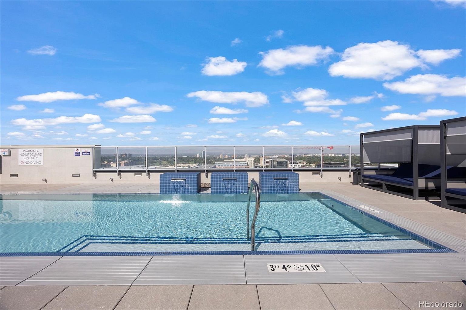 This image showcases a rooftop pool area with a modern design. The pool features a tiled edge and a shallow end, with a metal railing for access. In the background, there's a cityscape view and cabanas, suggesting a luxurious and relaxing amenity for residents.