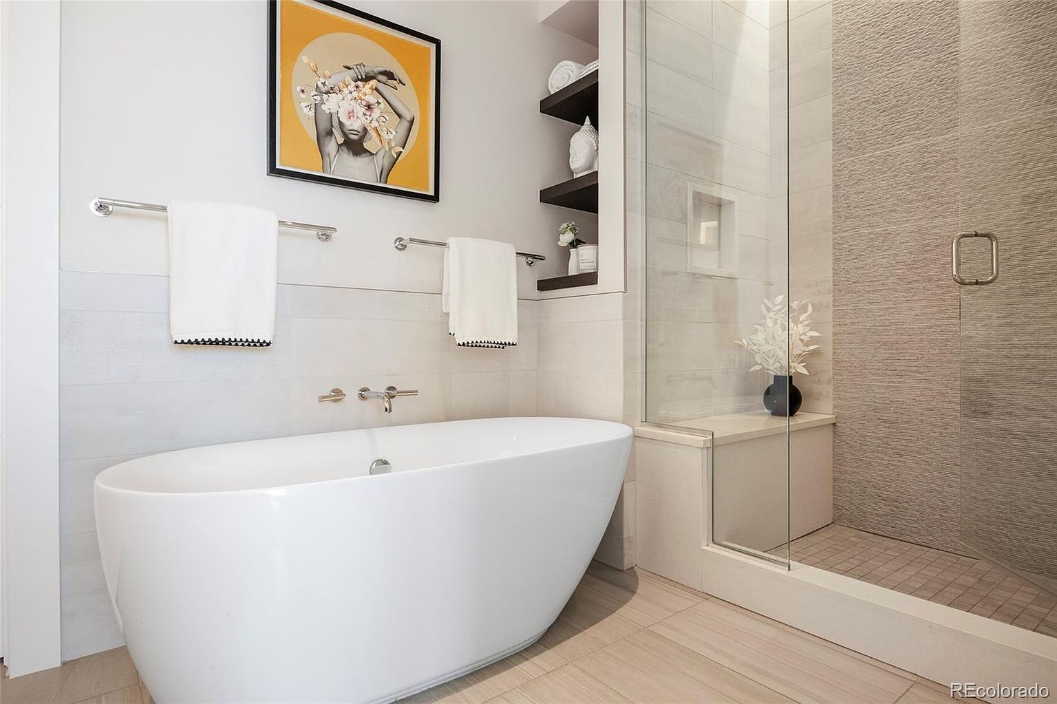 This is a primary bathroom featuring a freestanding white bathtub, a glass-enclosed shower with textured tile, and modern fixtures. The walls are a light neutral tone, complemented by dark wood shelving and a framed art piece. The overall impression is clean, luxurious, and spa-like.
