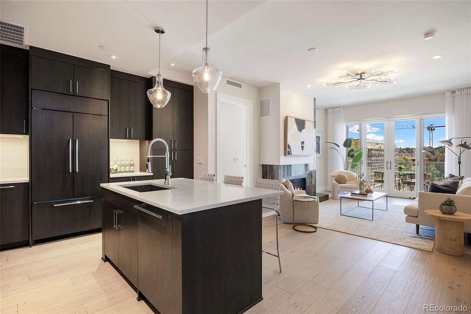 This interior shot showcases a modern kitchen seamlessly integrated with a living area. The kitchen features dark wood cabinetry, a white countertop island with a sink, and pendant lighting. The living area includes a fireplace, comfortable seating, and access to a balcony through sliding glass doors, creating an open and inviting space.