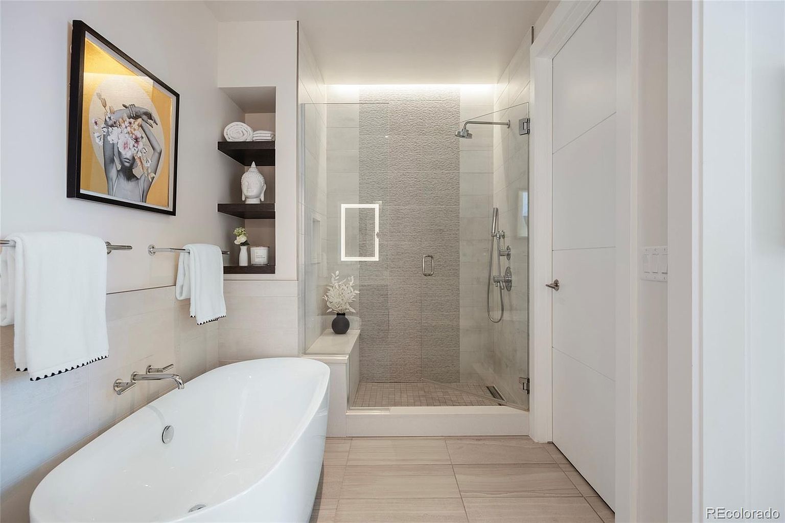 This is a bright and modern primary bathroom featuring a freestanding soaking tub, a glass-enclosed shower with a rainfall showerhead, and stylish shelving. The neutral color palette, combined with the artwork and fixtures, creates a spa-like atmosphere. The tile flooring and clean lines contribute to the bathroom's contemporary design.