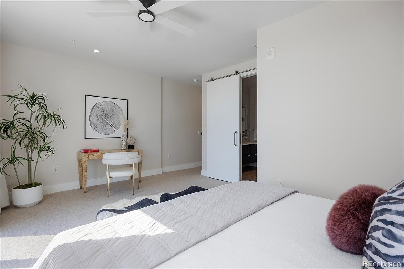 This is a bright and airy primary bedroom featuring neutral tones and modern decor. The room includes a desk area with a decorative plant and artwork, a sliding barn door leading to the bathroom, and a bed with stylish pillows and a quilted throw. The overall impression is clean, comfortable, and inviting.