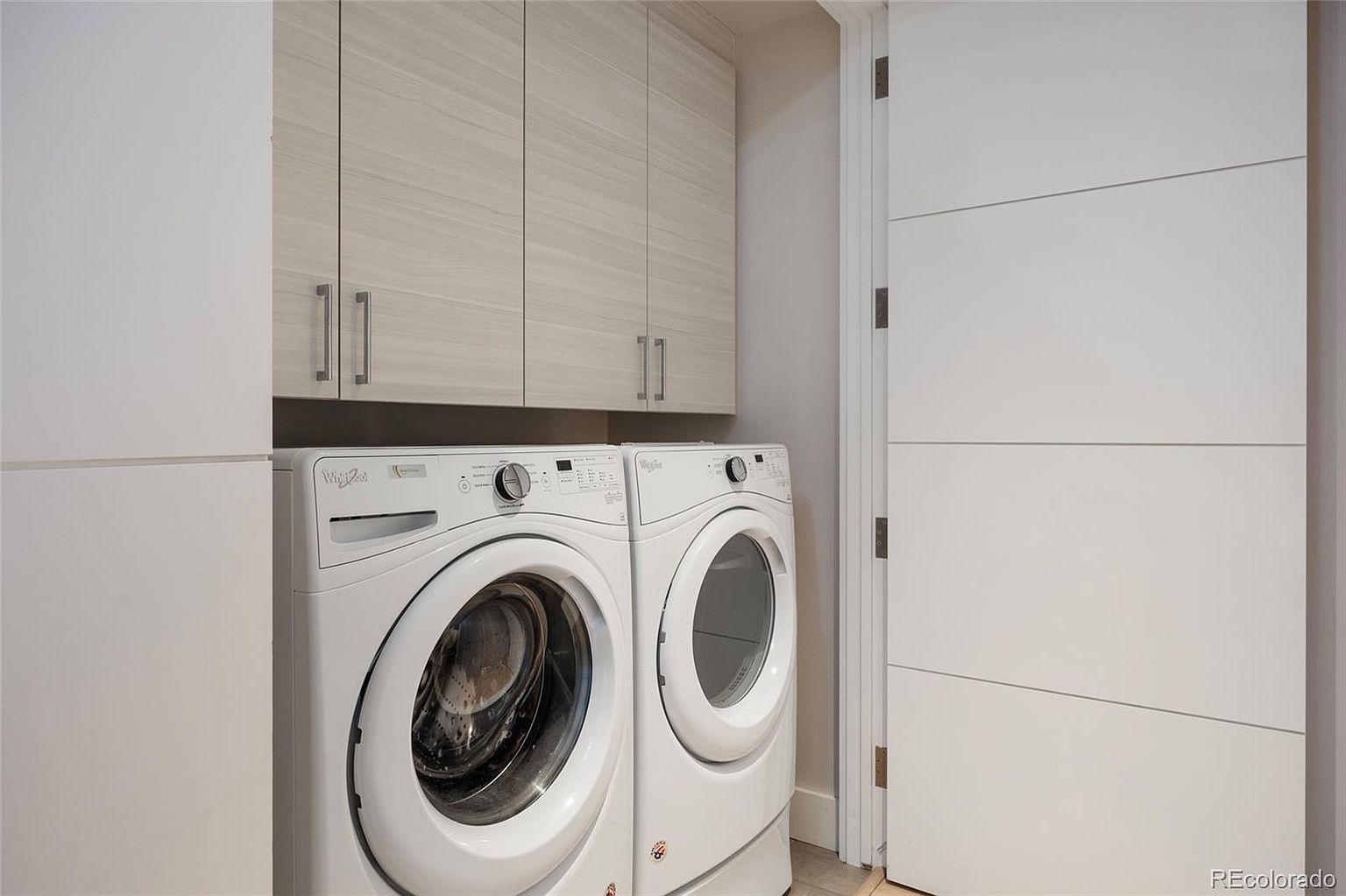 This is a well-organized laundry room featuring a front-loading washer and dryer set in white, complemented by light wood-tone cabinets above. The room has a clean and modern aesthetic, with a white door featuring horizontal paneling adding to the contemporary design. The space appears functional and efficient, ideal for a modern home.