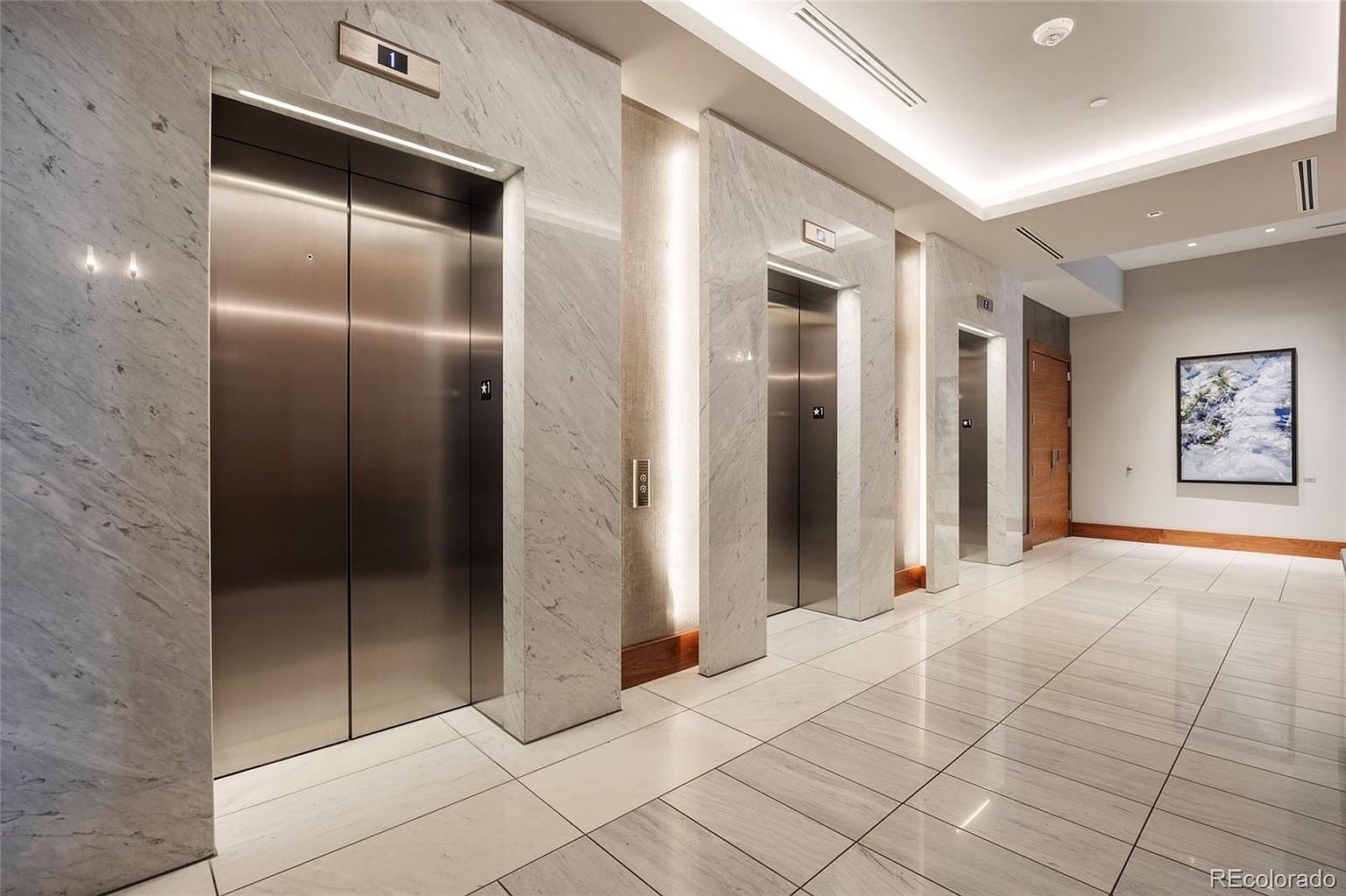 This interior shot showcases a modern hallway with three elevator banks, each framed by marble pillars with integrated lighting. The floor is tiled in a matching marble pattern, creating a cohesive and luxurious aesthetic. A framed piece of art hangs on the wall at the end of the hallway, adding a touch of sophistication.