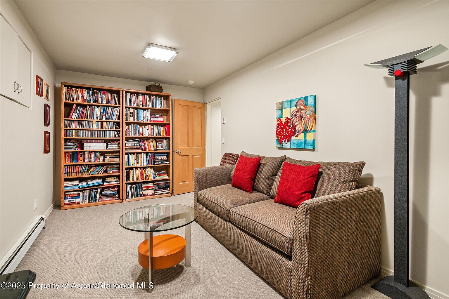 This interior shot showcases a cozy living room featuring a brown sofa with red pillows, a glass-topped coffee table, and two full bookshelves. An abstract painting hangs on the wall above the sofa, and a unique floor lamp stands in the corner. The room is carpeted and appears well-lit, creating a comfortable and inviting atmosphere.