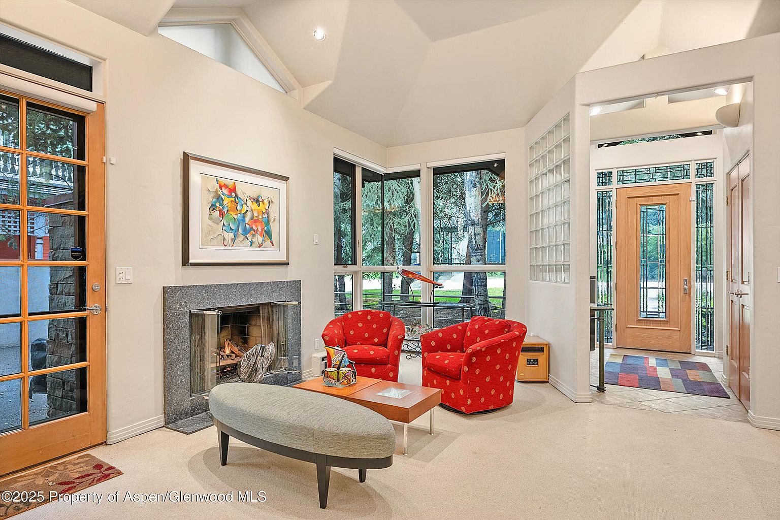 This is an interior shot of a living room featuring a fireplace, two red armchairs, and a bench. The room has large windows offering natural light and views of the outdoors. The style is comfortable and inviting, with a mix of traditional and modern elements.
