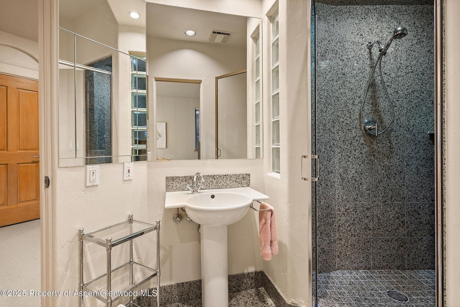 This is a bathroom featuring a pedestal sink with a granite countertop, a large mirror, and a glass-enclosed shower with gray granite-like tiling. The bathroom has a neutral color palette with white walls and gray flooring, creating a clean and modern look. A chrome shelving unit adds storage space, and the overall impression is one of functionality and style.