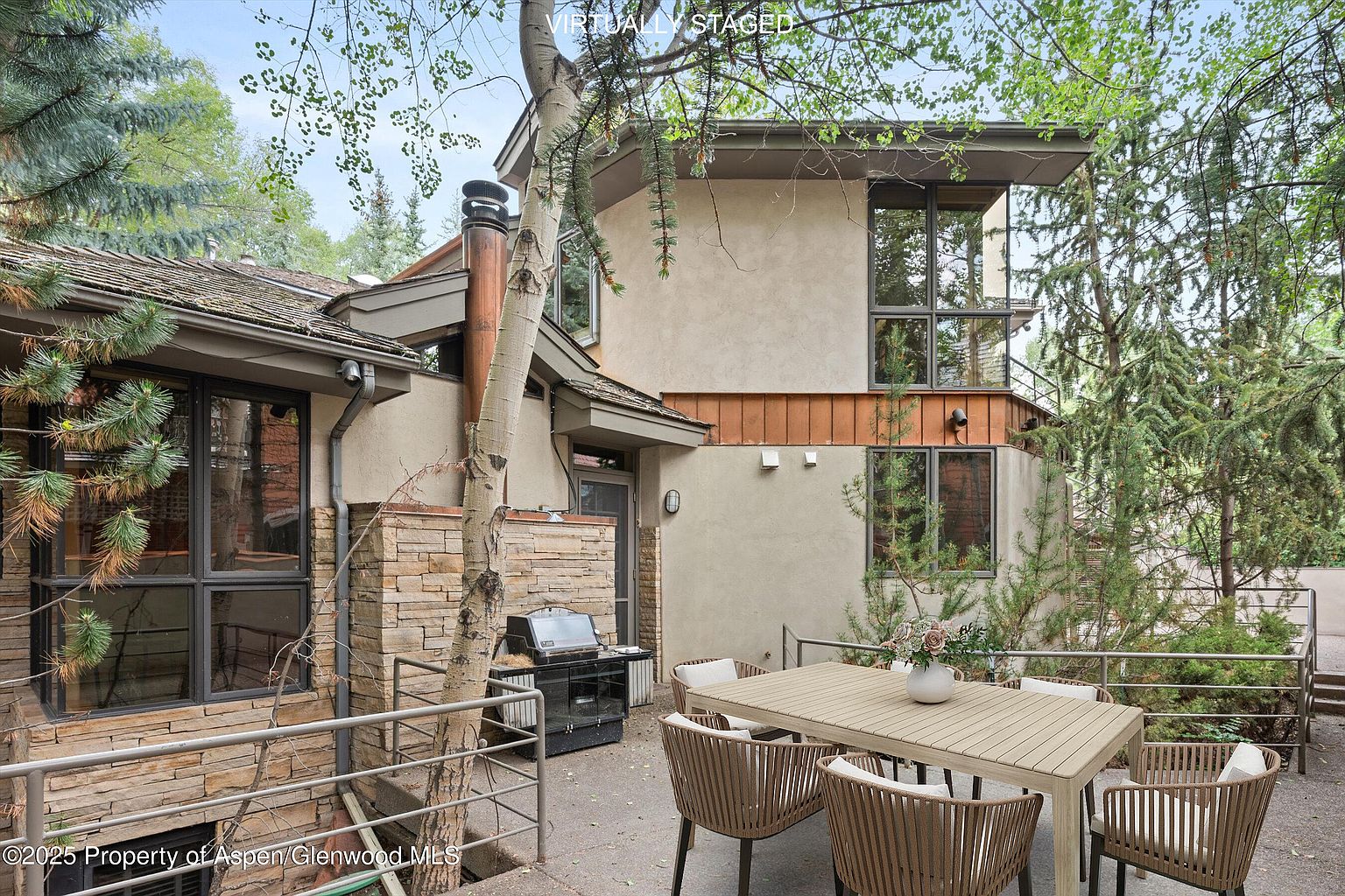 This image showcases an inviting outdoor patio area of a home. The patio features a dining table with chairs, a grill, and a stone accent wall, creating a cozy and functional space for outdoor entertaining. The surrounding trees and landscaping provide privacy and a natural backdrop.