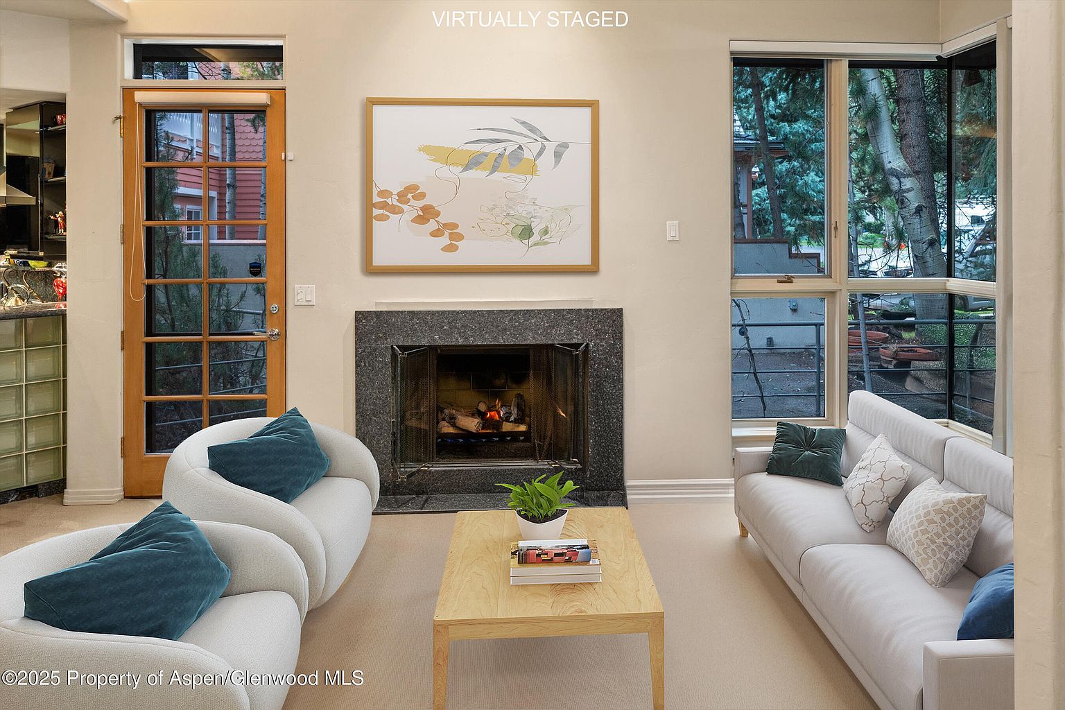 This is an interior shot of a living room, virtually staged. The room features a fireplace with a framed artwork above it, two white curved sofas with teal pillows, and a light wood coffee table. Large windows provide natural light and views of the outdoors, creating a bright and inviting atmosphere.