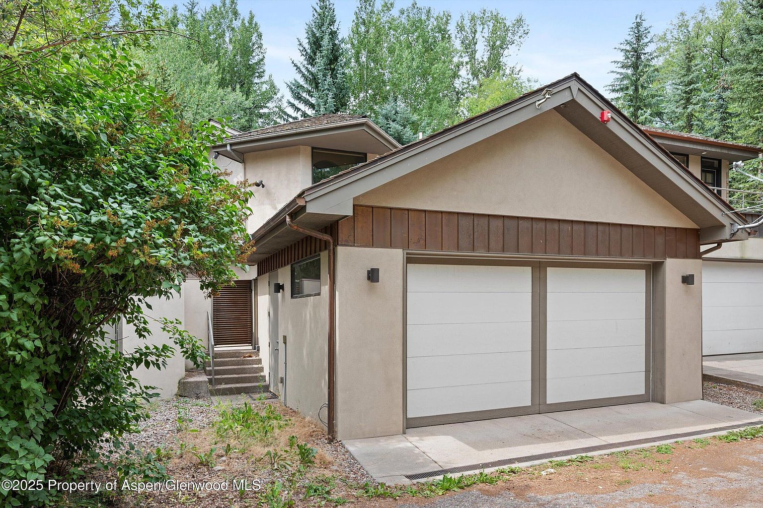 This image showcases the exterior of a home, focusing on the garage. The garage features two white doors with a neutral trim, complemented by a section of brown wood paneling above. The exterior walls are a light beige stucco, and the overall style is modern and well-maintained, with mature trees in the background adding to the property's appeal.