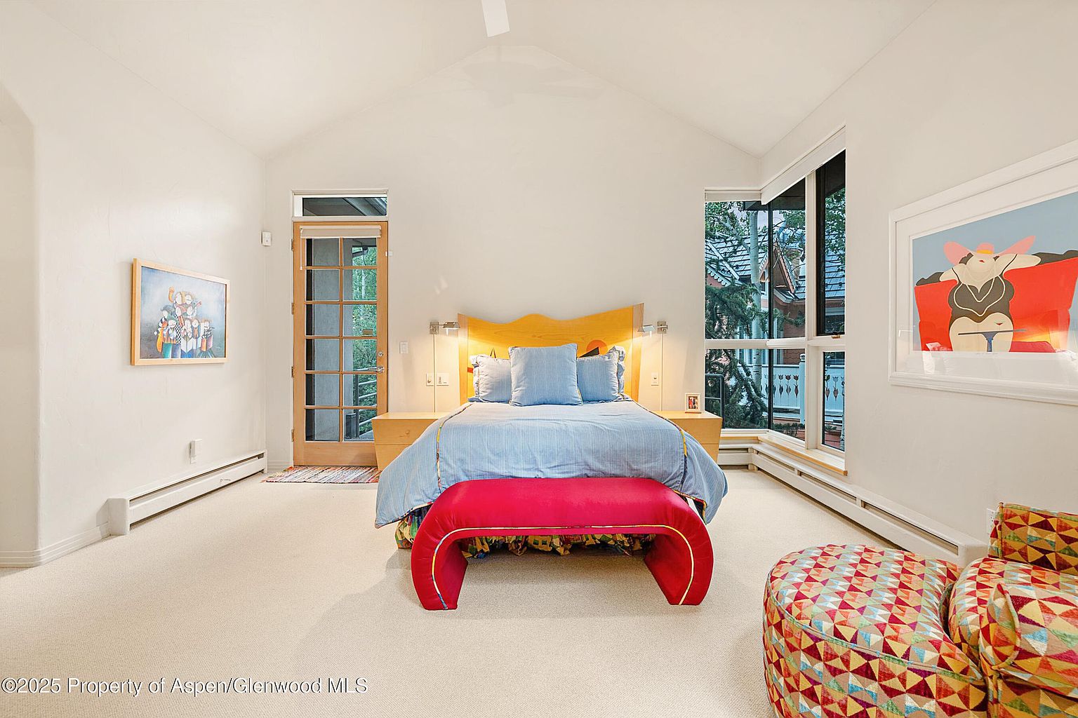 This is a bright and airy primary bedroom featuring a unique headboard and a red bench at the foot of the bed. The room is well-lit with natural light from the windows and complemented by modern light fixtures. The overall impression is a comfortable and stylish space.