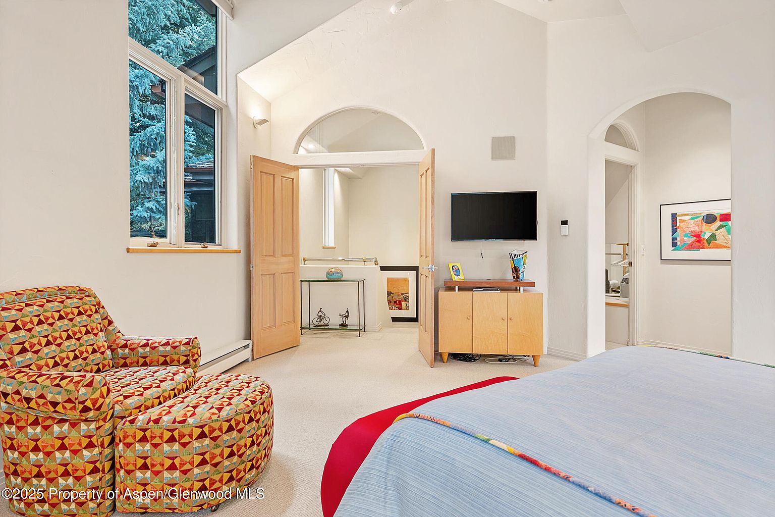 This is a bright and airy primary bedroom featuring an armchair with a matching ottoman, a television mounted on the wall, and an arched doorway leading to another room. The room is painted in neutral tones, and the bed is partially visible in the foreground with a blue and red cover. The overall impression is one of comfort and relaxation.