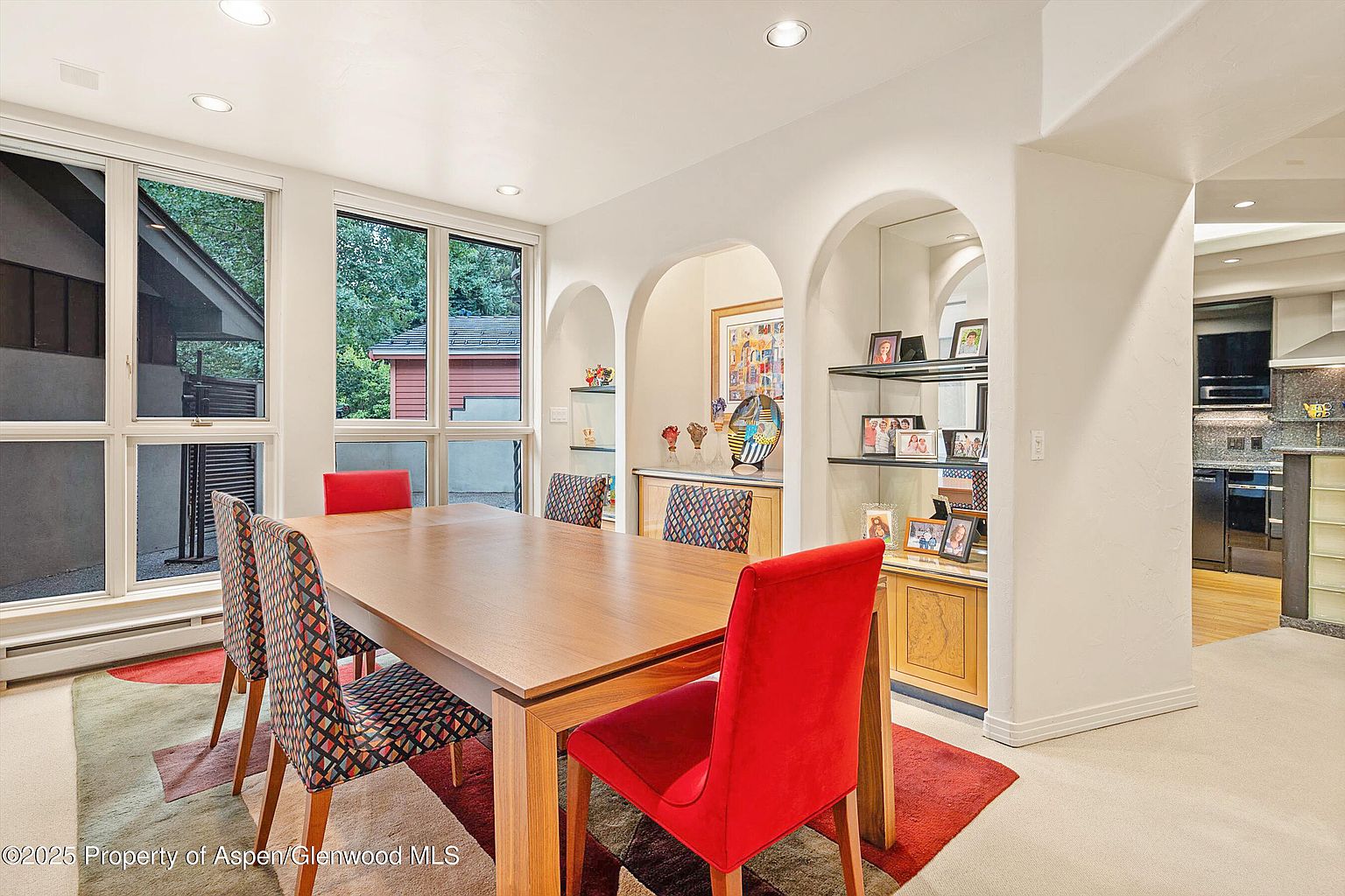 This is an interior shot of a dining room featuring a large wooden dining table surrounded by chairs with red and patterned upholstery. Natural light floods the room through large windows, and built-in shelving with arched openings adds architectural interest. The room is decorated with artwork and personal items, creating a warm and inviting atmosphere.
