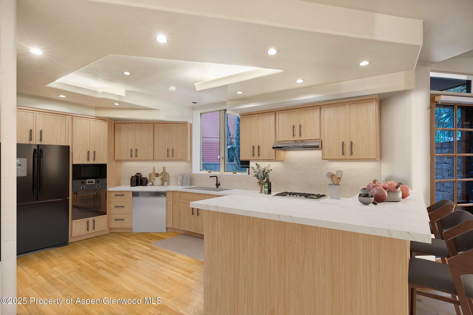 This is a well-lit kitchen featuring light wood cabinetry with modern hardware, a black refrigerator, and a white countertop island with seating. The kitchen is equipped with stainless steel appliances and has a window providing natural light. The overall impression is a clean and functional space.