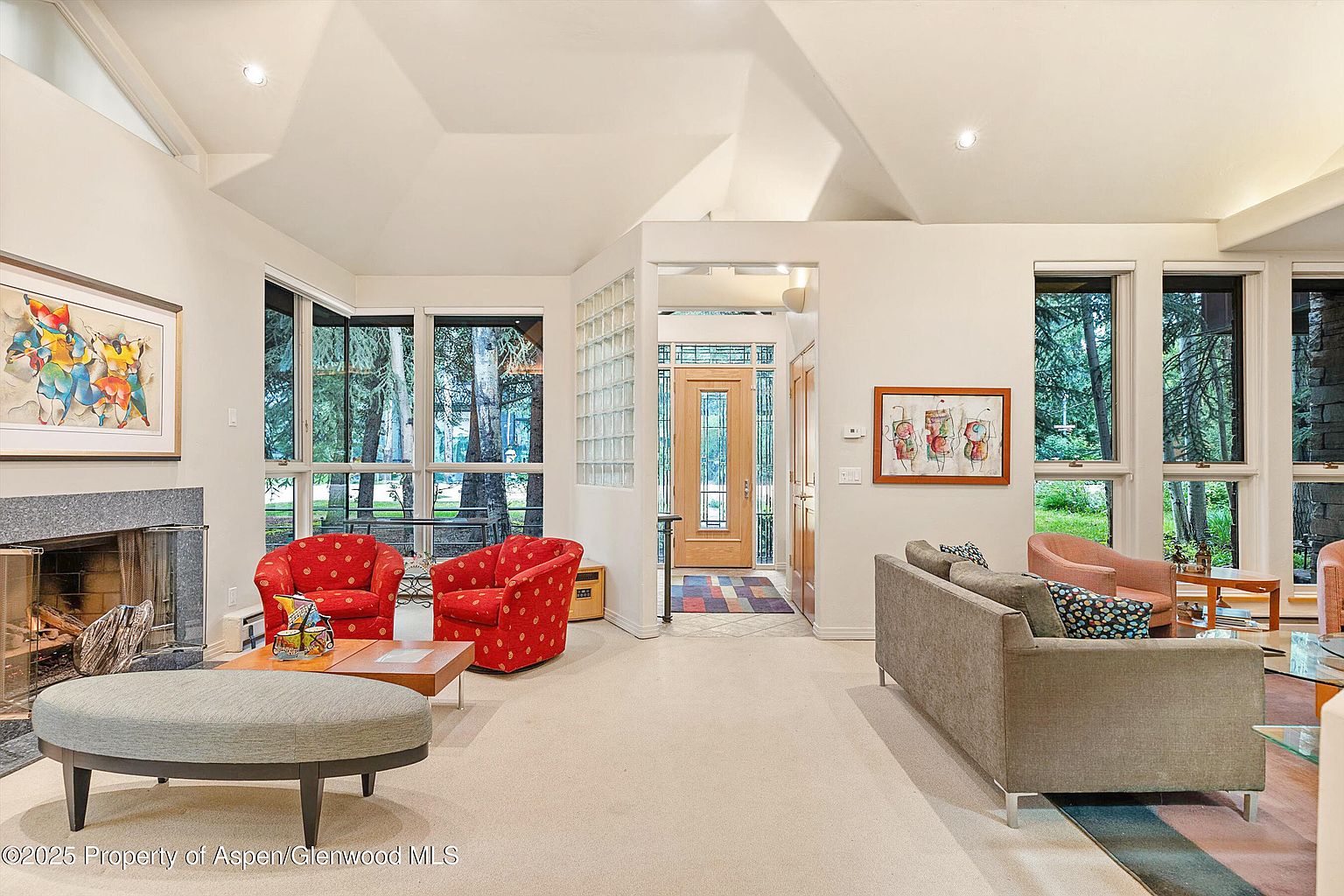 This is an interior shot of a living room featuring a fireplace, two red armchairs, and a gray sofa. Large windows provide natural light and views of the surrounding trees. The room has a neutral color palette with pops of color from the furniture and artwork, creating a warm and inviting atmosphere.