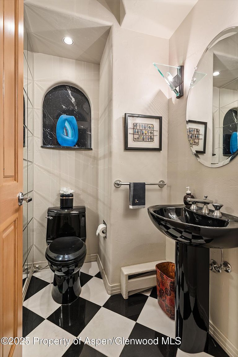 This is a stylish guest bathroom featuring a black toilet and pedestal sink set against a black and white checkered floor. The walls are a neutral tone, and the room includes unique architectural details such as a recessed niche with a blue decorative piece and a modern glass sconce. The overall impression is sophisticated and contemporary.