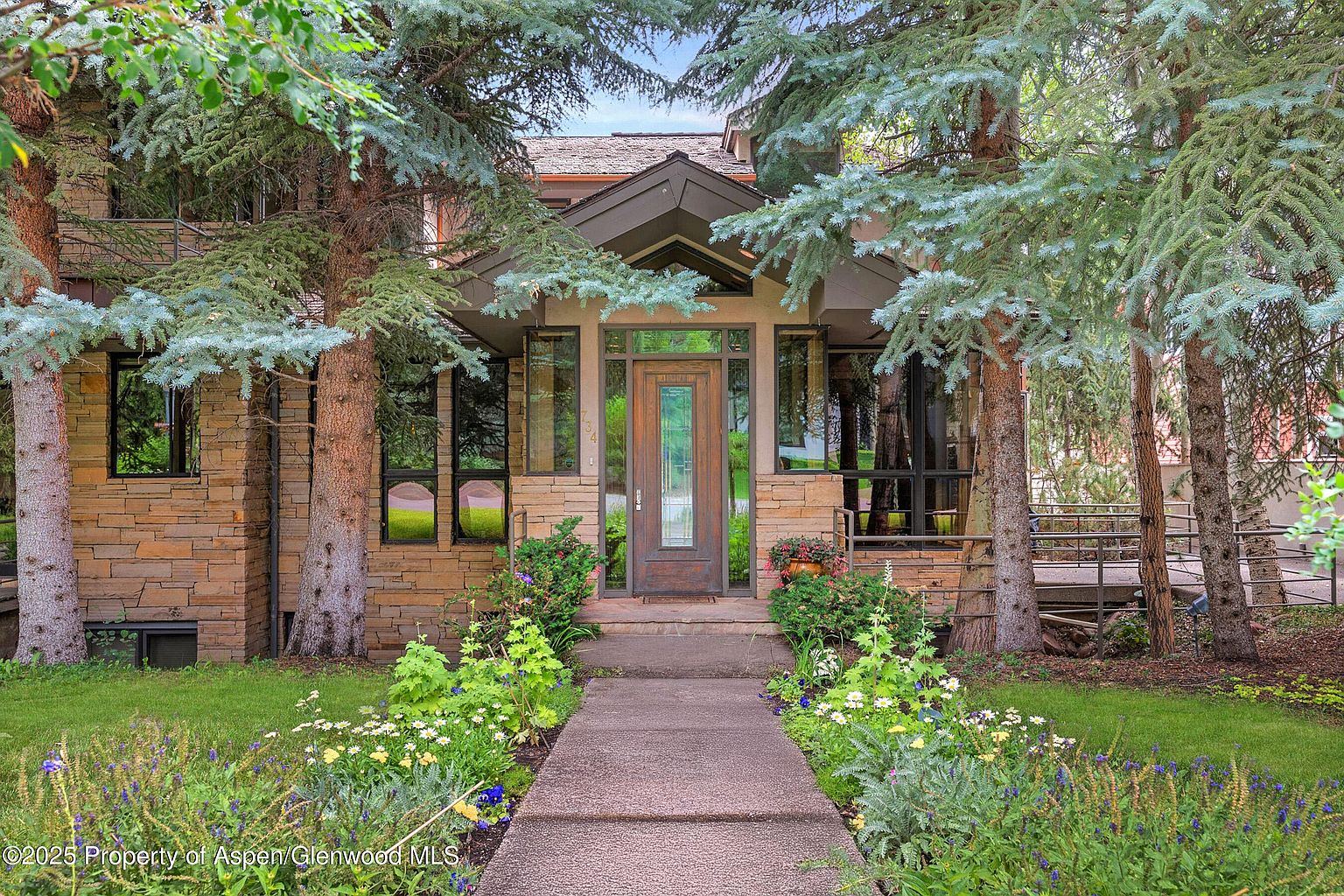 This image showcases the inviting entryway of a home, framed by mature evergreen trees. A stone pathway leads to a wooden front door, flanked by windows and stone facade. The landscaping features a variety of flowers and greenery, adding to the curb appeal.
