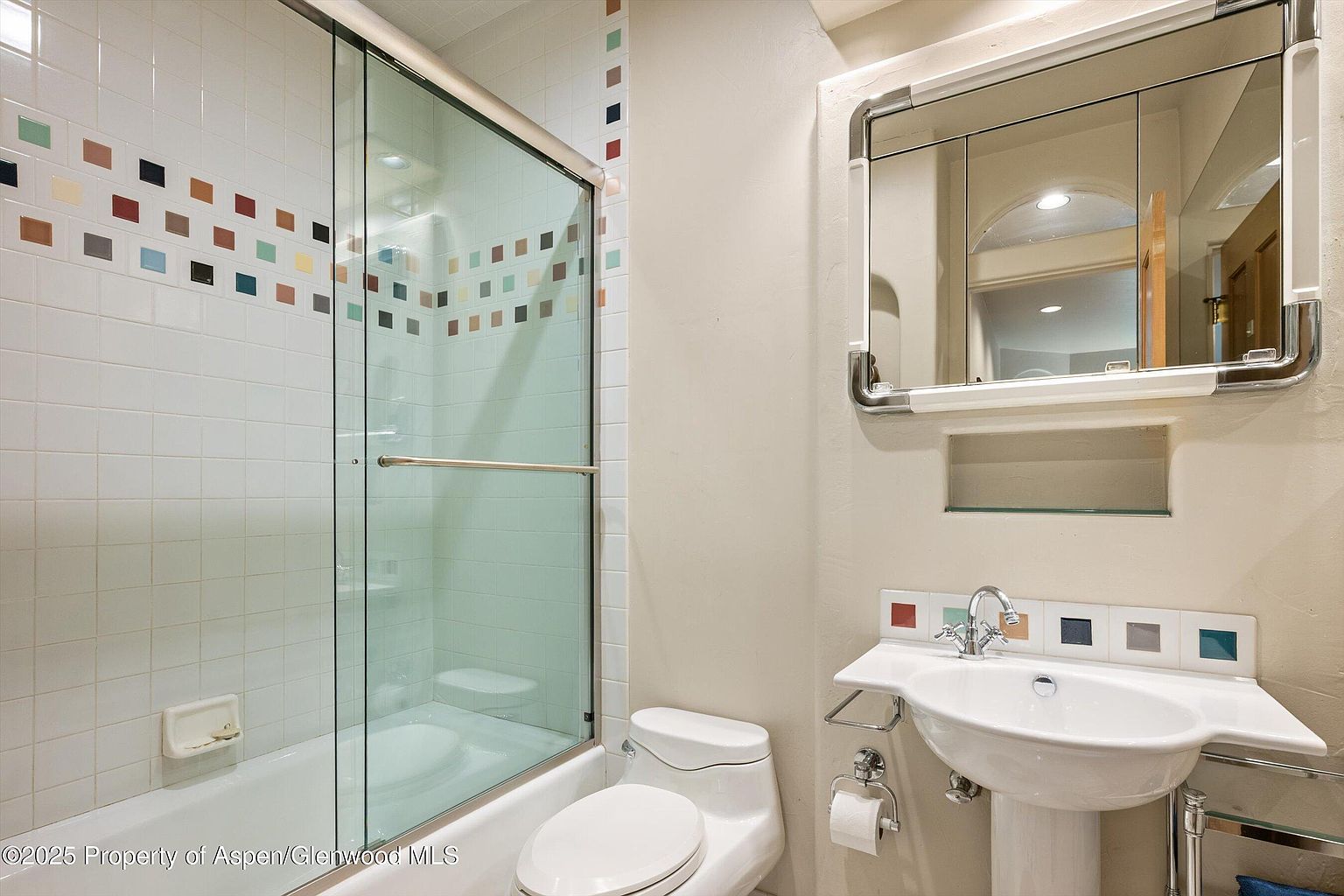 This is a bathroom featuring a bathtub with a glass shower door, a toilet, and a pedestal sink. The walls are tiled with white squares accented by a row of colorful square tiles. Above the sink is a mirrored medicine cabinet with chrome trim, and the overall style is clean and functional.