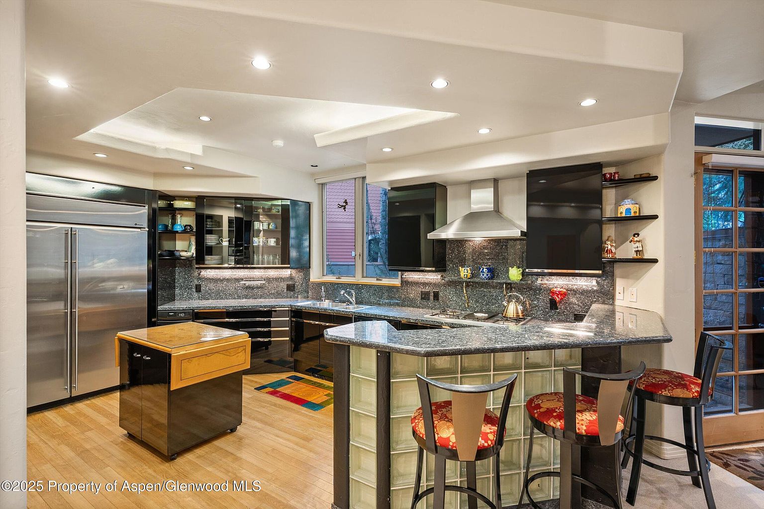 This is a well-lit kitchen featuring dark cabinetry with glass fronts, granite countertops, and stainless steel appliances, including a large refrigerator. A kitchen island with a wooden top and a breakfast bar with glass block accents provide additional workspace and seating. The flooring is light-colored hardwood, and recessed lighting illuminates the space, creating a modern and functional culinary environment.