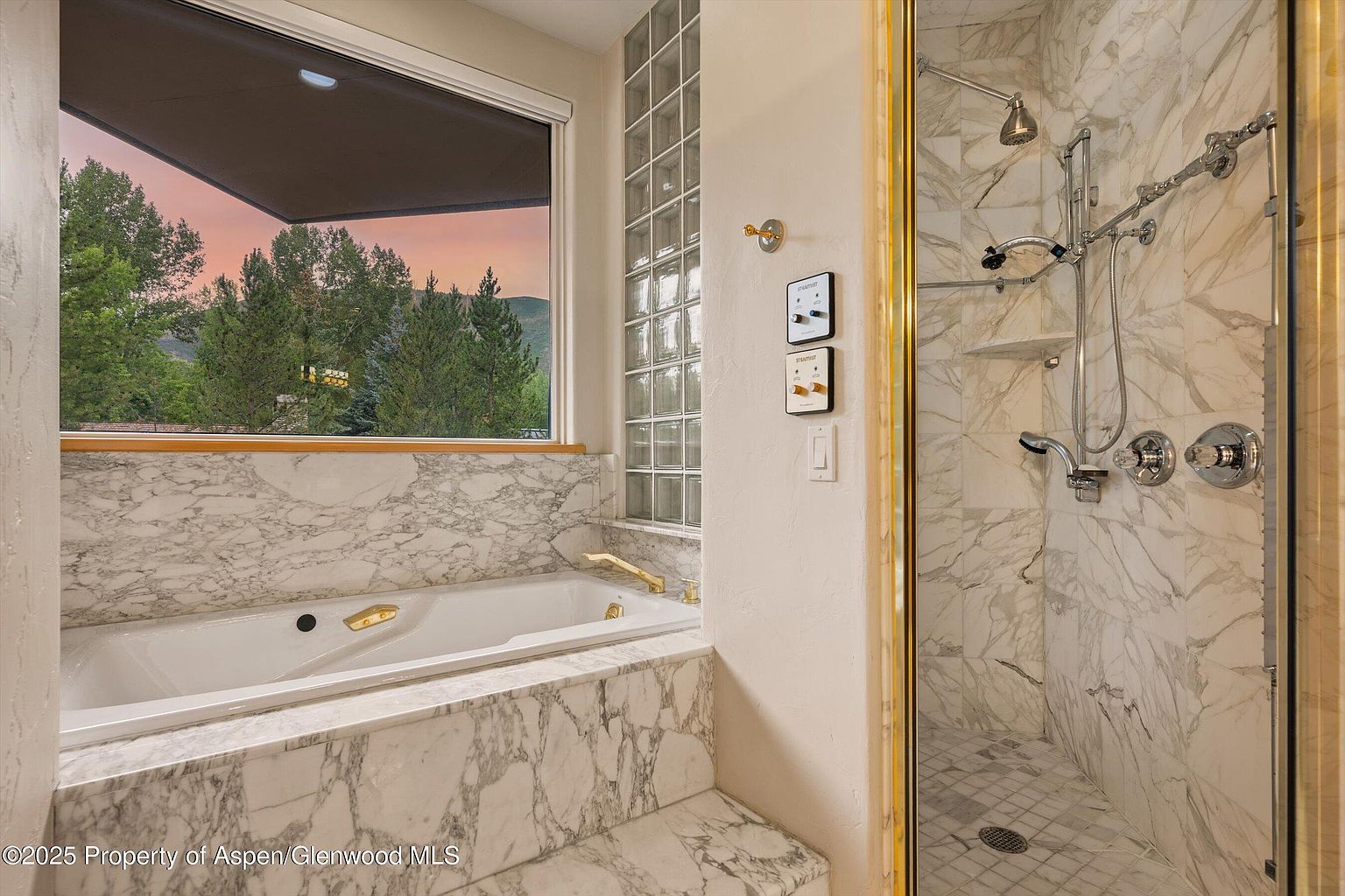 This primary bathroom features a luxurious marble-clad bathtub and a separate shower with marble walls and a glass door. A large window offers a view of the outdoors, bringing natural light into the space. The bathroom exudes a sense of elegance and relaxation, perfect for a spa-like experience.