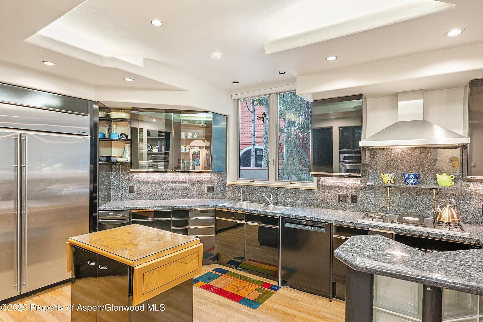 This is a well-lit kitchen featuring black cabinetry, granite countertops, and stainless steel appliances, including a large refrigerator. A central island with a wooden top provides additional workspace, and a window above the sink offers natural light. The kitchen has a modern and sophisticated aesthetic.
