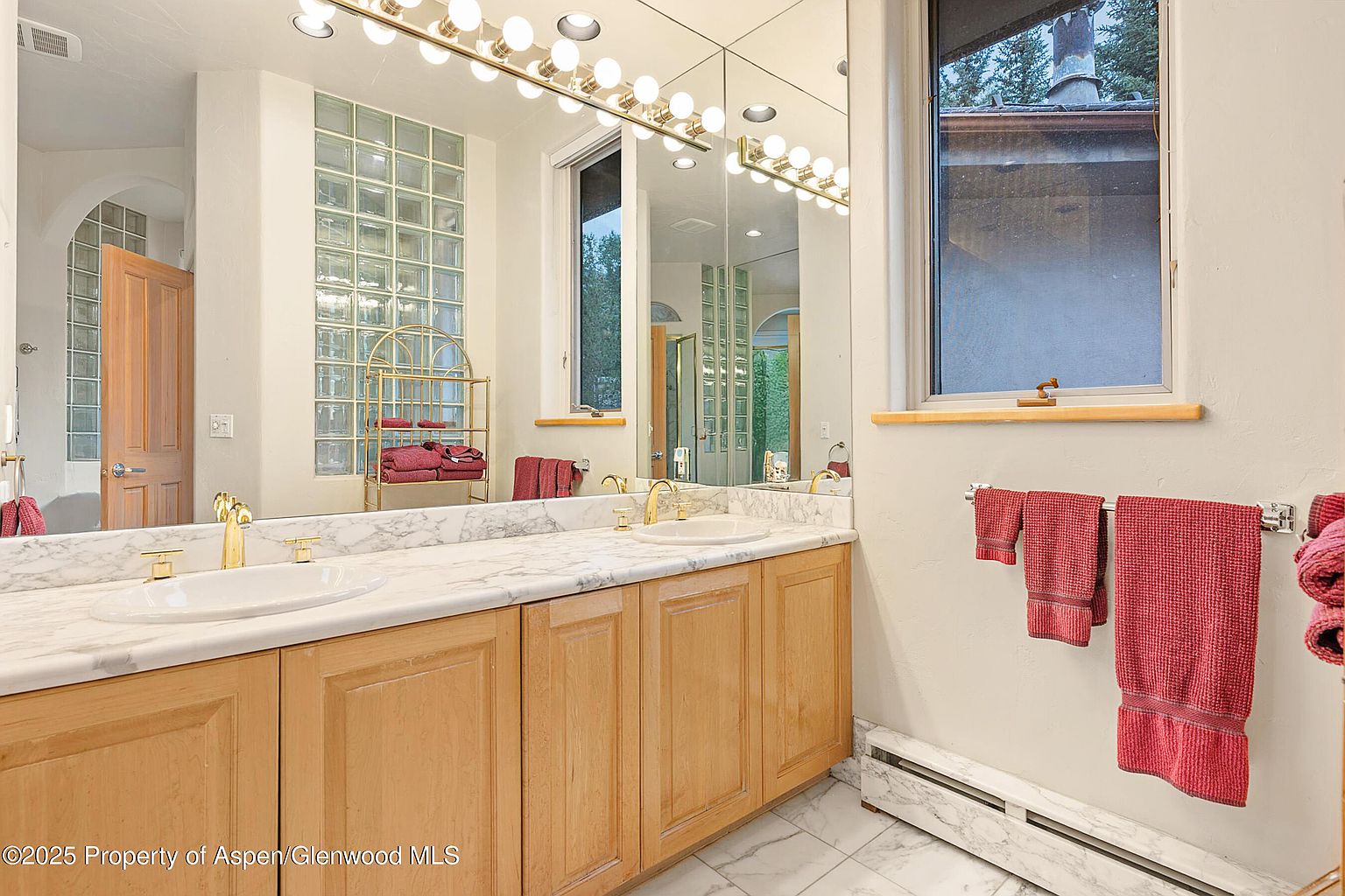 This is a well-lit primary bathroom featuring a double vanity with a marble countertop and light wood cabinetry. A large mirror with vanity lighting spans the length of the counter, reflecting a glass block window and a view outside. Red towels add a pop of color, and the overall impression is clean and functional.