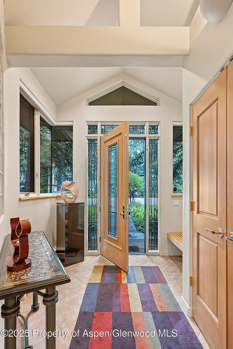This is an inviting entryway featuring a partially open wooden door with glass panels, flanked by windows that offer a view of the outdoors. A colorful striped rug leads into the space, complemented by a decorative table and a modern sculpture. The area is well-lit and appears spacious, creating a welcoming first impression.