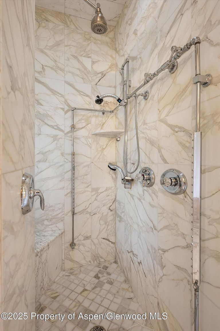 This is a luxurious primary bathroom featuring a spacious walk-in shower. The shower is adorned with elegant marble tiling and equipped with multiple shower heads and chrome fixtures. A built-in bench provides seating, and the overall design exudes a spa-like ambiance.