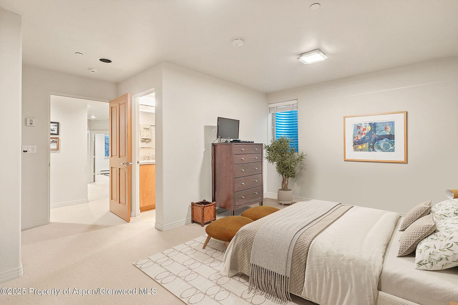 This is a well-lit primary bedroom featuring a neutral color palette and a calming atmosphere. The room includes a bed with soft linens, a dresser with a television, and a small seating area. A window with blue blinds provides natural light, and artwork adorns the walls, creating a serene and inviting space.
