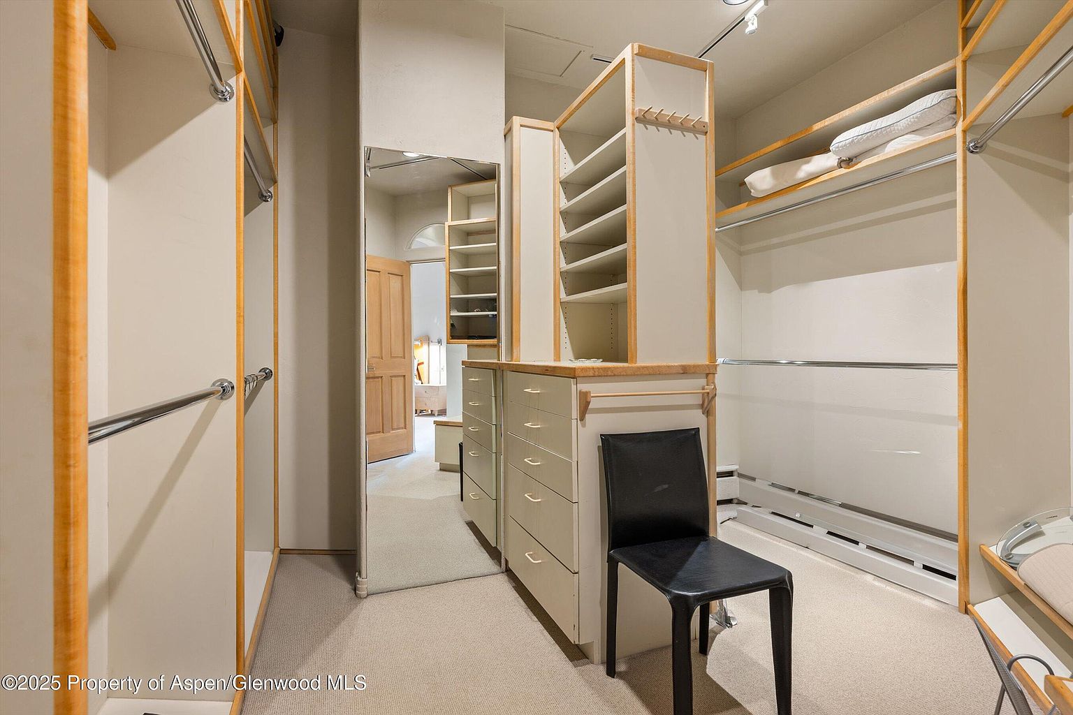 This is a well-organized walk-in closet featuring custom shelving and drawers with light wood trim. A black chair sits in the center, and a mirror reflects the closet's depth. The closet is carpeted and well-lit, offering ample storage space.