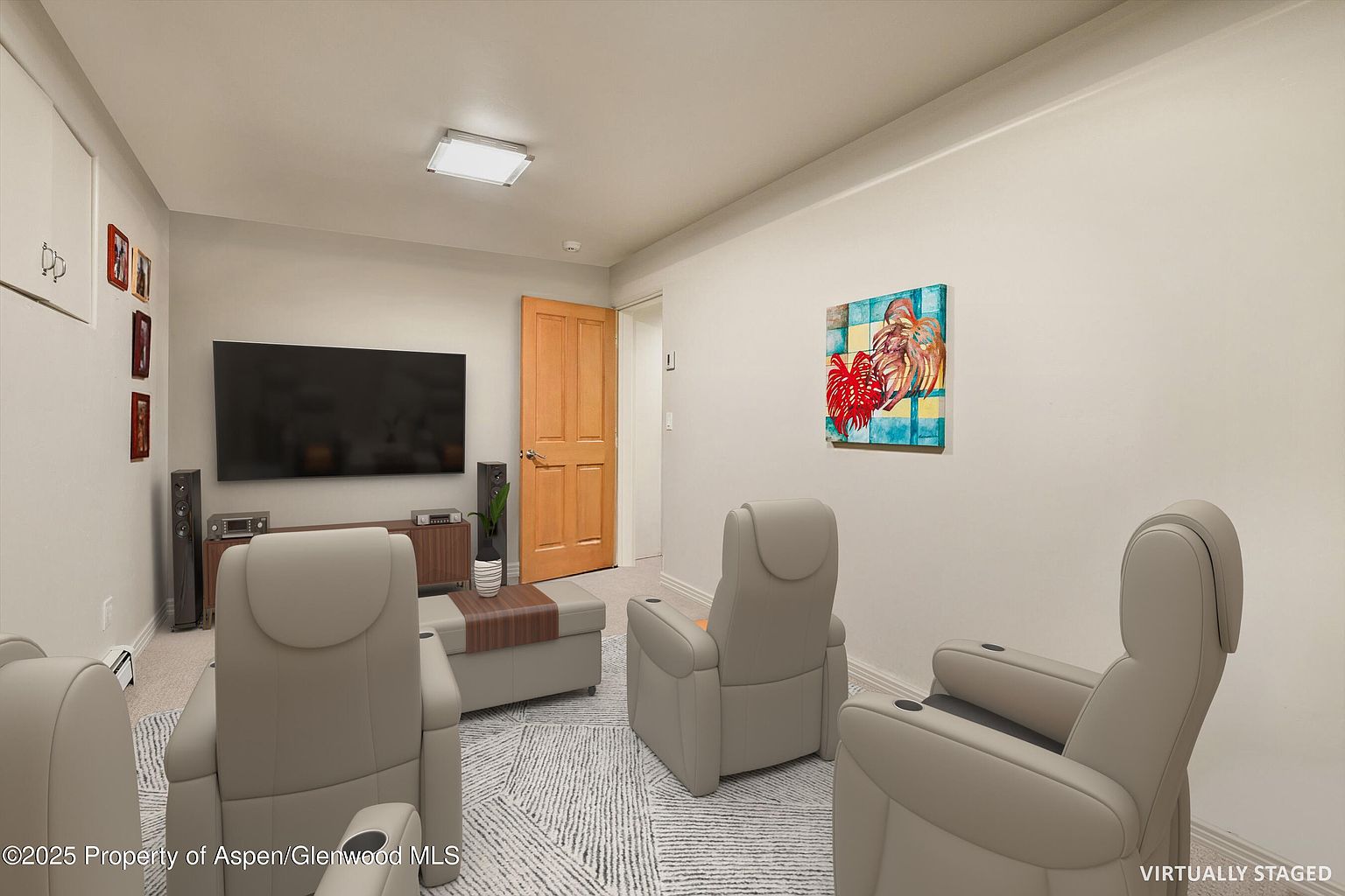 This is an interior shot of a living room, likely a home theater setup. The room features several reclining chairs arranged around a large television. A wooden door is visible on the right side of the image, and a piece of artwork hangs on the wall. The room is carpeted and has neutral-colored walls, creating a cozy and inviting atmosphere.