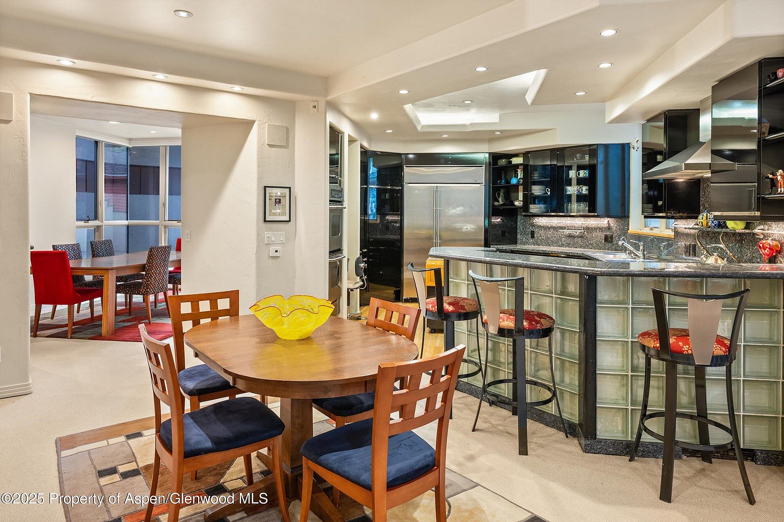 This interior shot showcases a modern kitchen and dining area. The kitchen features sleek black cabinetry, stainless steel appliances, and a unique glass block bar. The dining area includes a round wooden table with blue cushioned chairs, complemented by a patterned rug, creating a warm and inviting space.