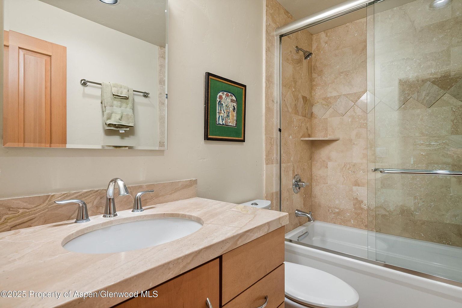 This is a well-lit bathroom featuring a vanity with a light-colored stone countertop and wooden cabinetry. A mirror hangs above the sink, reflecting a towel rack with neatly folded towels. The bathroom also includes a bathtub with a glass shower door, surrounded by beige marble tiles, creating a clean and inviting space.